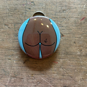Bottle opener keychain featuring a design of a brown butt with an anchor tattoo on a blue background on top of a wooden surface. 