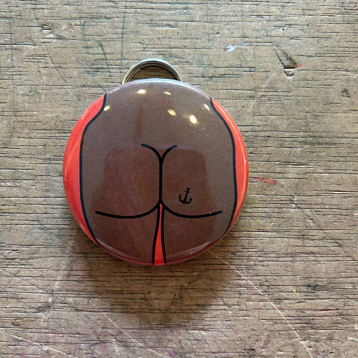 Bottle opener keychain featuring a design of a brown butt with an anchor tattoo on an orange background on top of a wooden surface. 