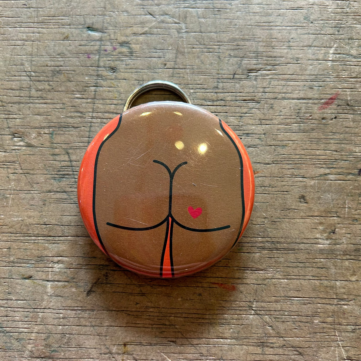 Bottle opener keychain featuring a design of a brown butt with a heart tattoo on an orange background on top of a wooden surface. 