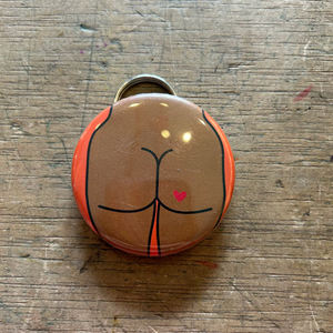 Bottle opener keychain featuring a design of a brown butt with a heart tattoo on an orange background on top of a wooden surface. 