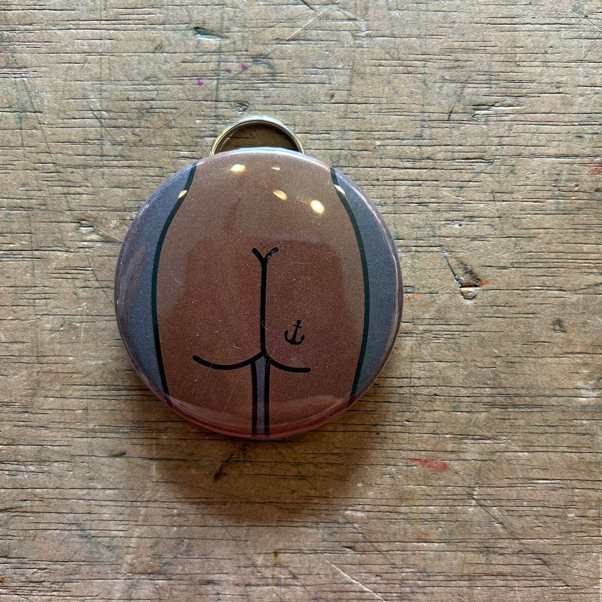 Bottle opener keychain featuring a design of a brown butt with an anchor tattoo on gray background on top of a wooden surface. 
