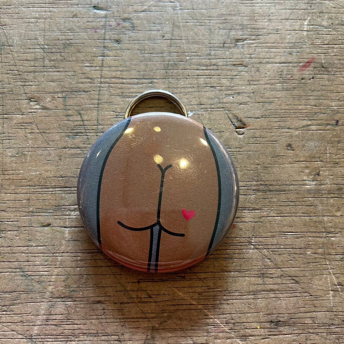 Bottle opener keychain featuring a design of a brown butt with a heart tattoo on a gray background on top of a wooden surface. 
