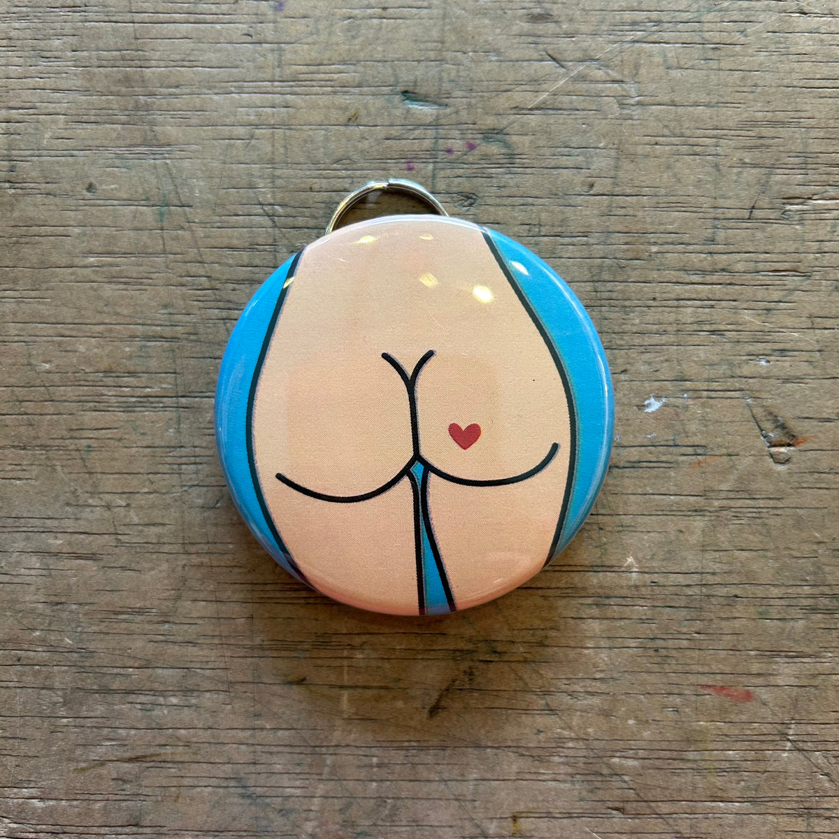 Bottle opener keychain featuring a design of a light colored butt with a red heart tattoo on a blue background on top of a wooden surface. 