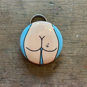 Bottle opener keychain featuring a design of a light colored butt with an anchor tattoo against a blue background on top of a wooden surface. 