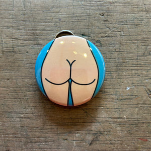 Bottle opener keychain featuring a design of a light colored butt on a blue background on top of a wooden surface. 