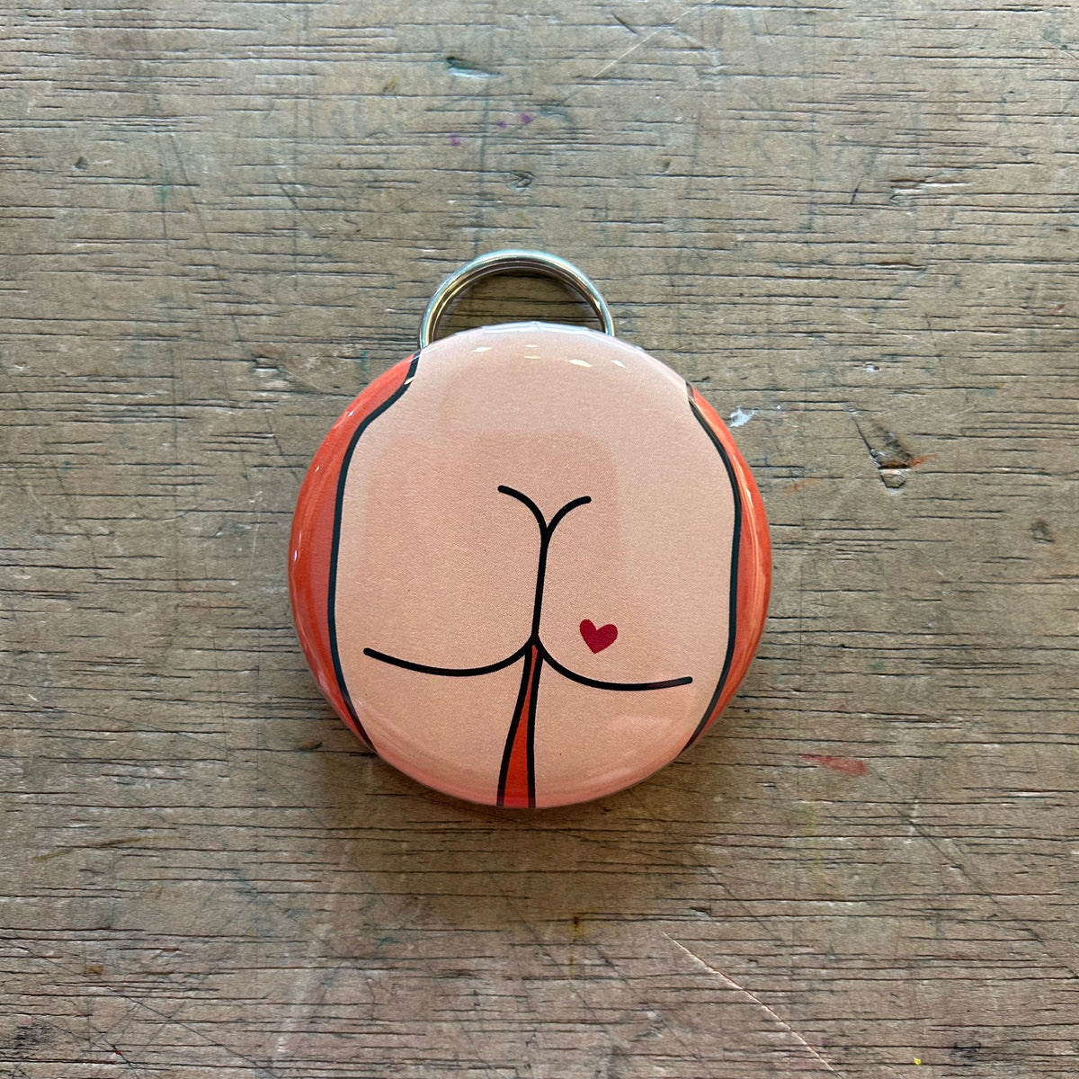 Bottle opener keychain featuring a light colored butt with a red heart tattoo on an orange background on top of a wooden surface. 