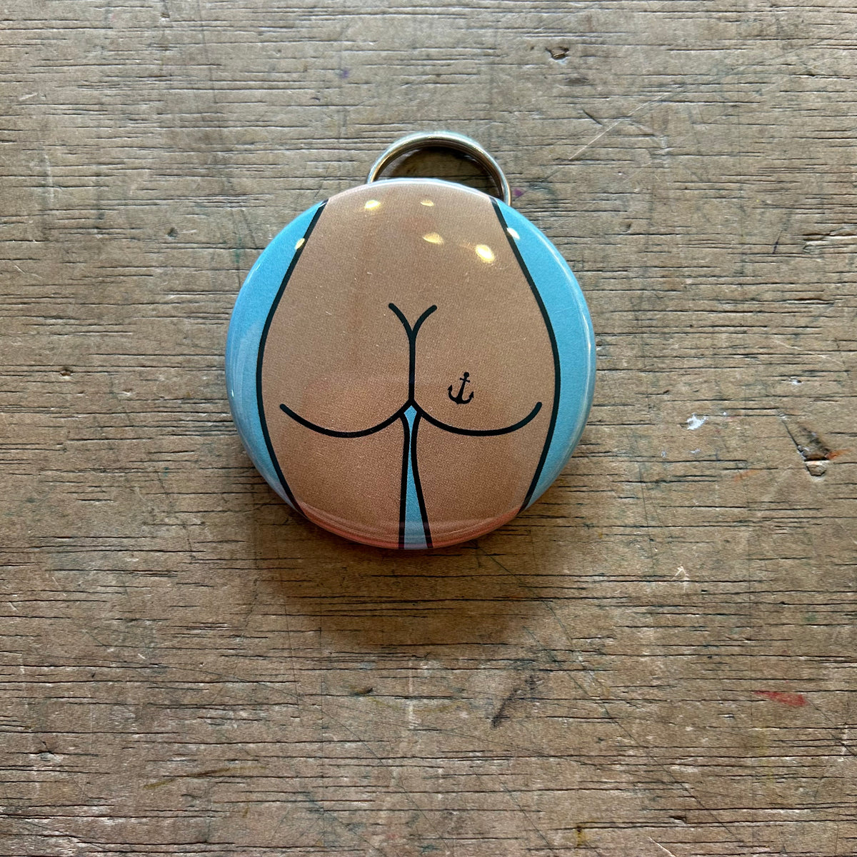 Bottle opener keychain featuring a design of a tan butt with an anchor tattoo on a blue background on top of a wooden surface. 