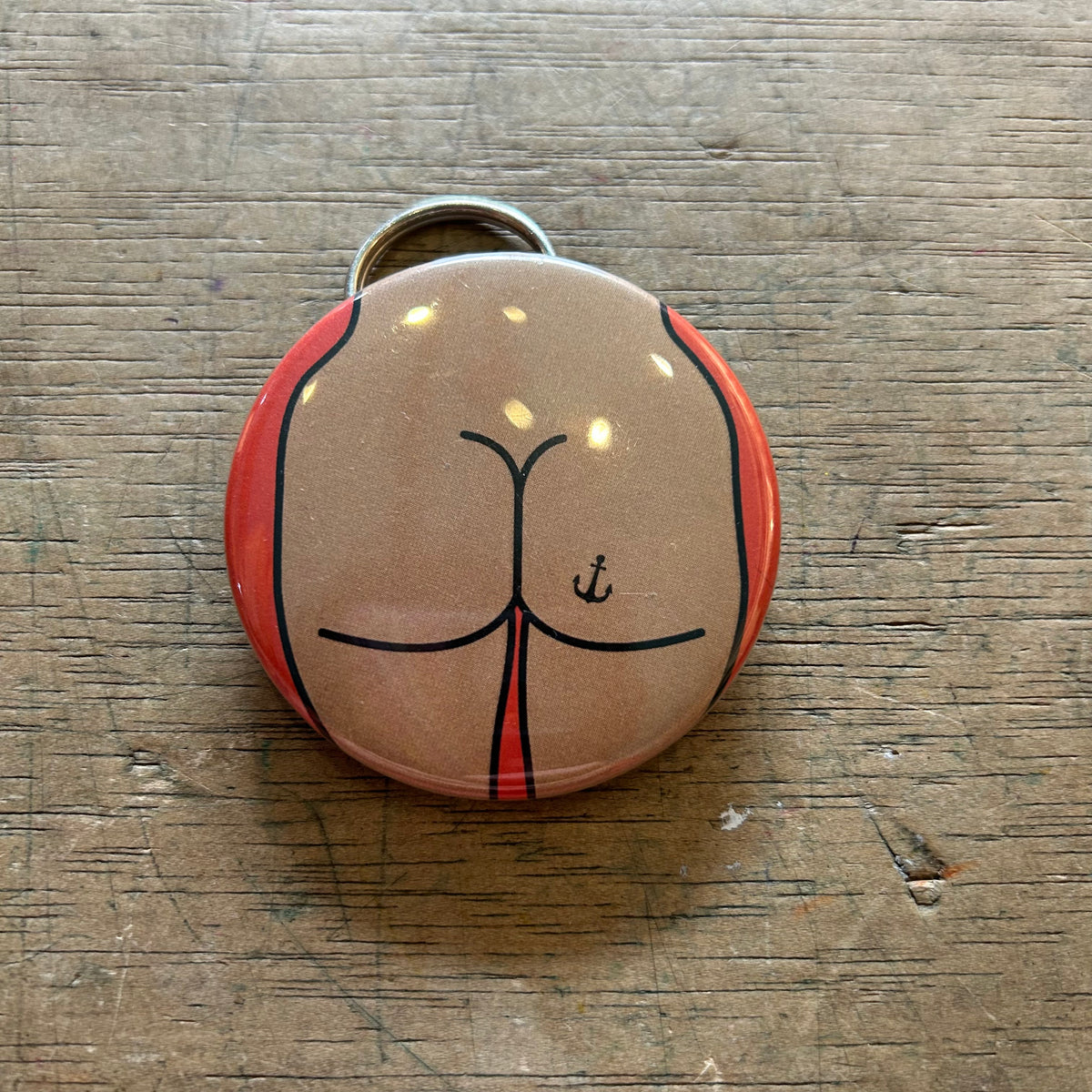 Bottle opener keychain featuring a design of a tan butt with an anchor tattoo against an orange background on top of a wooden surface. 