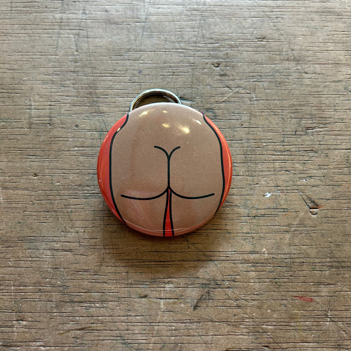 Bottle opener keychain featuring a design of a tan butt on an orange background on top of a wooden surface. 