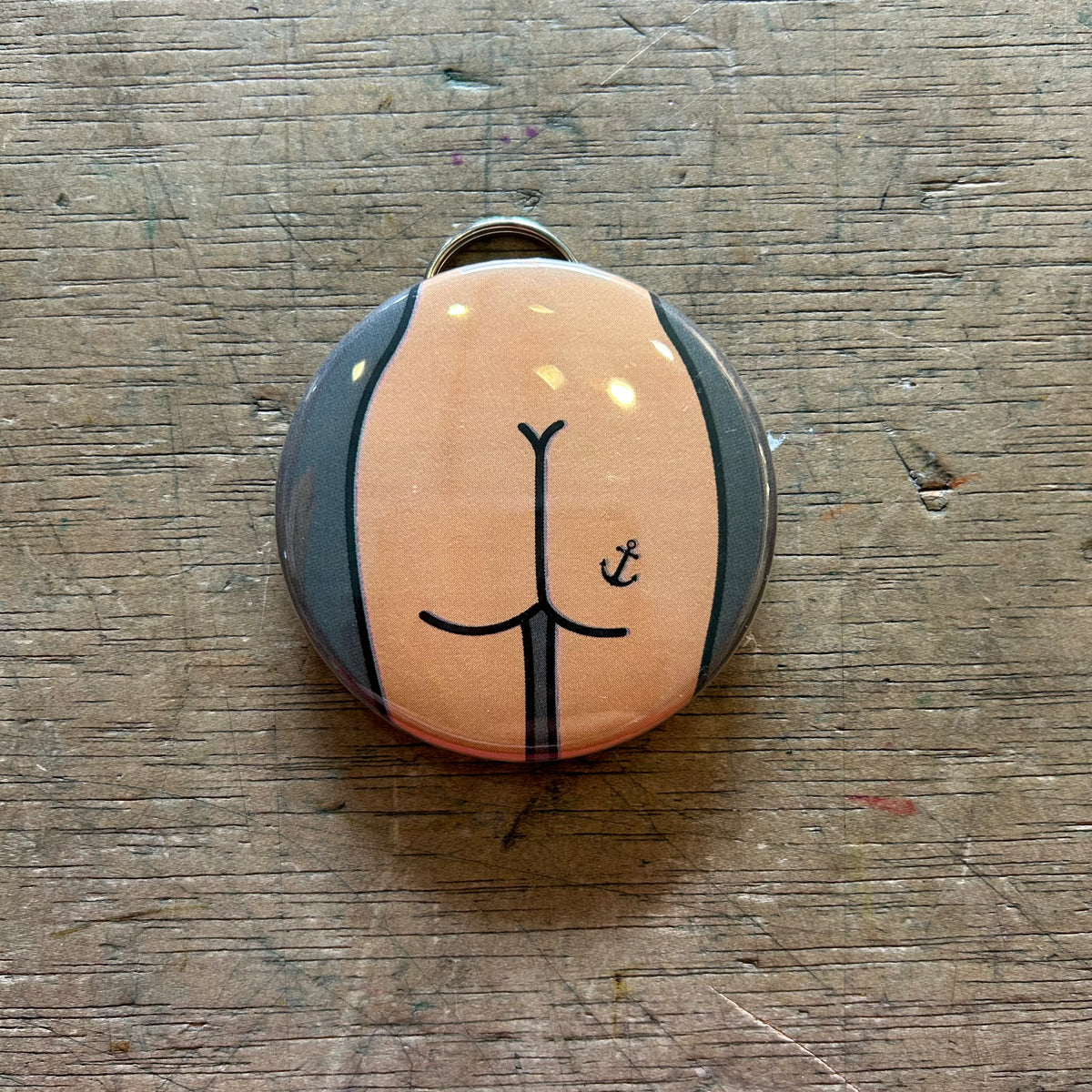 Bottle opener keychain featuring a tan butt with an anchor tattoo against a gray background on top of a wooden surface. 