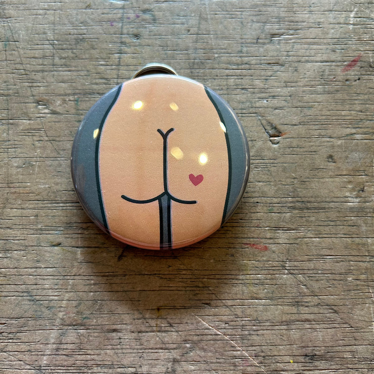 Bottle opener keychain featuring a design of a tan butt with a red heart tattoo on a gray background on top of a wooden surface. 