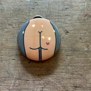 Bottle opener keychain featuring a design of a tan butt with a red heart tattoo on a gray background on top of a wooden surface. 