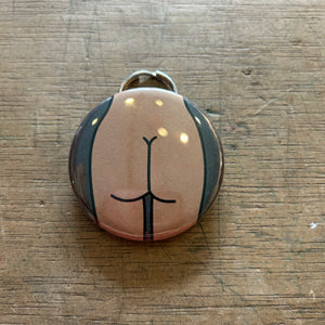 Bottle opener keychain featuring a design of a tan colored butt on a gray background on top of a wooden surface. 