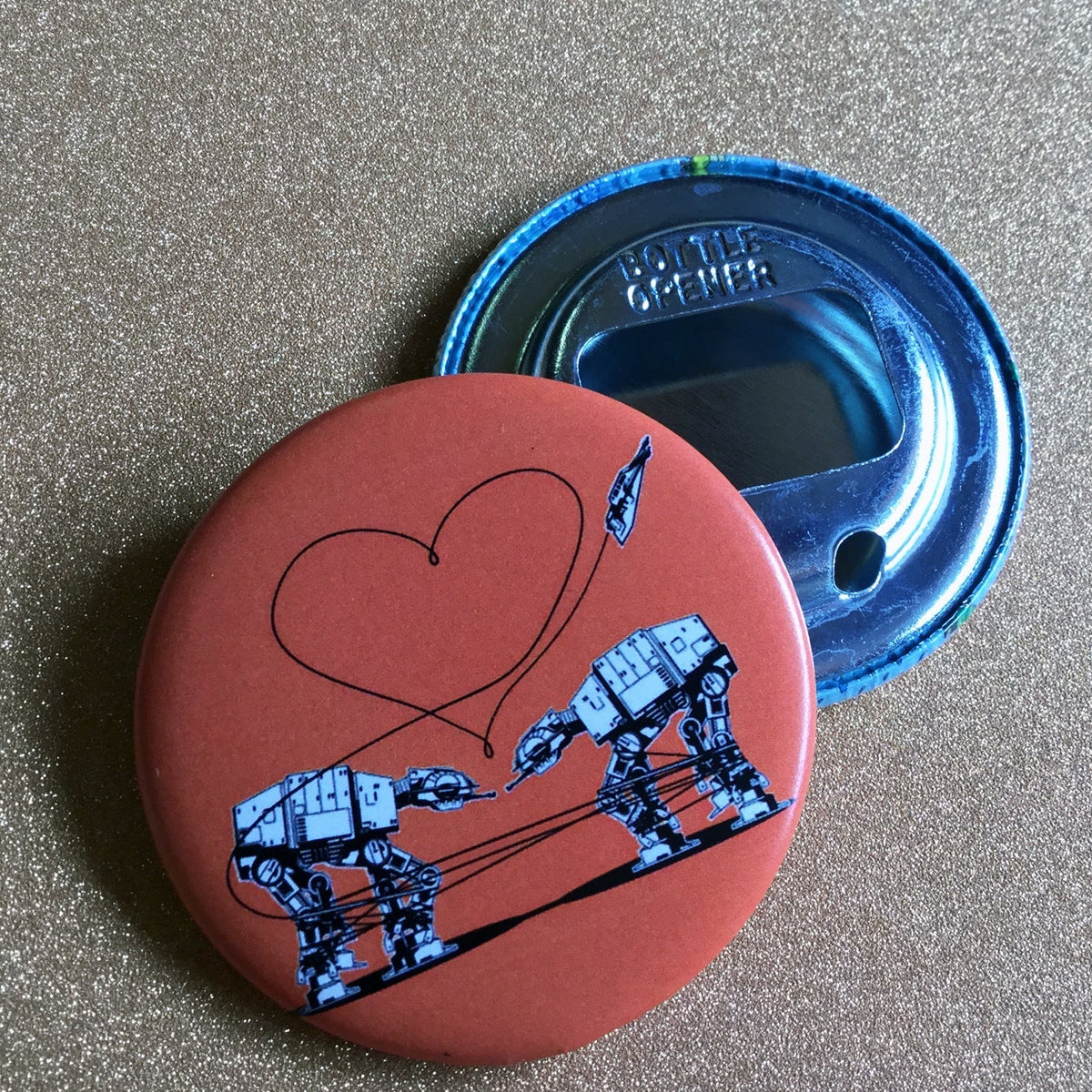 Round bottle opener keychain featuring the Love AT-AT First Sight design in orange against a gold glittery background. 