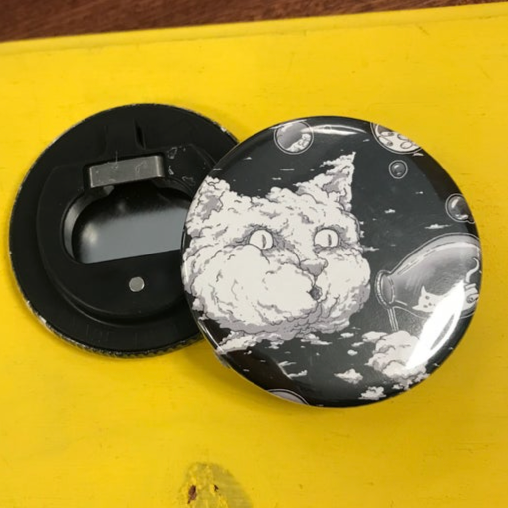 A round bottle opener with a black and white design of a cat blowing bubbles on top of a bright yellow surface. 