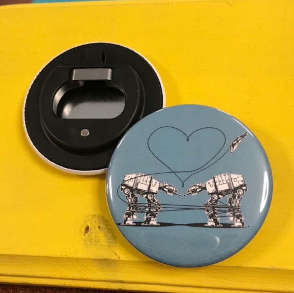 Round bottle opener magnet featuring the Love AT-AT First Sight design on a blue background on top of a yellow surface. 