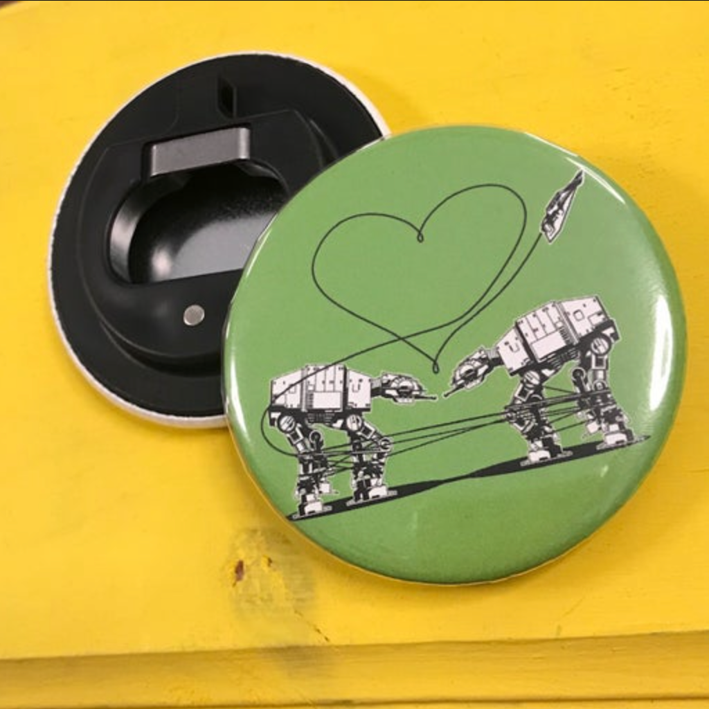 Round bottle opener magnet featuring the Love AT-AT First Sight design on a green background on top of a yellow surface. 