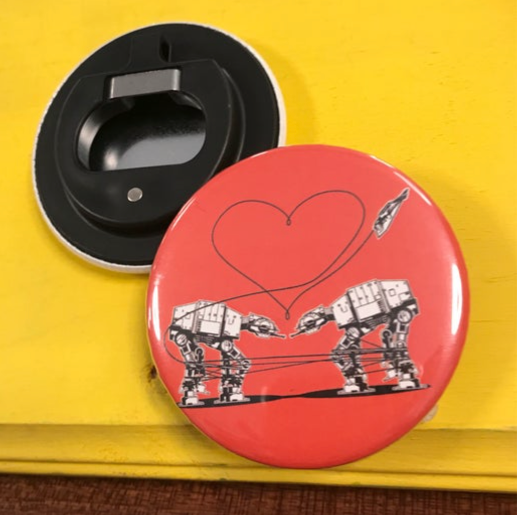 Round bottle opener magnet featuring the Love AT-AT First Sight design on an orange background on top of a yellow surface. 