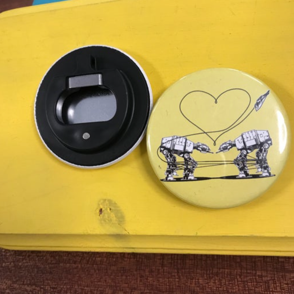 Round yellow bottle opener magnet featuring the Love AT-AT First Sight design on top of a yellow wooden surface. 