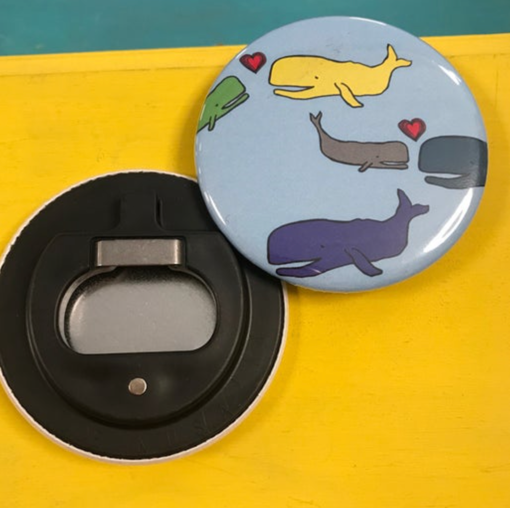 Round bottle opener magnet featuring whales of different colors against a light blue background. 