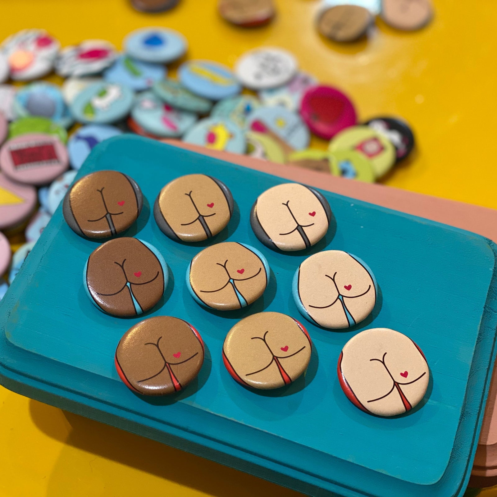 Set of buttons with cartoon butt designs on a blue tray against a colorful button background.