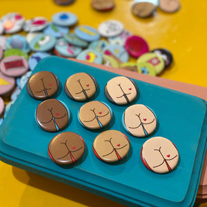 Set of buttons with cartoon butt designs on a blue tray against a colorful button background.