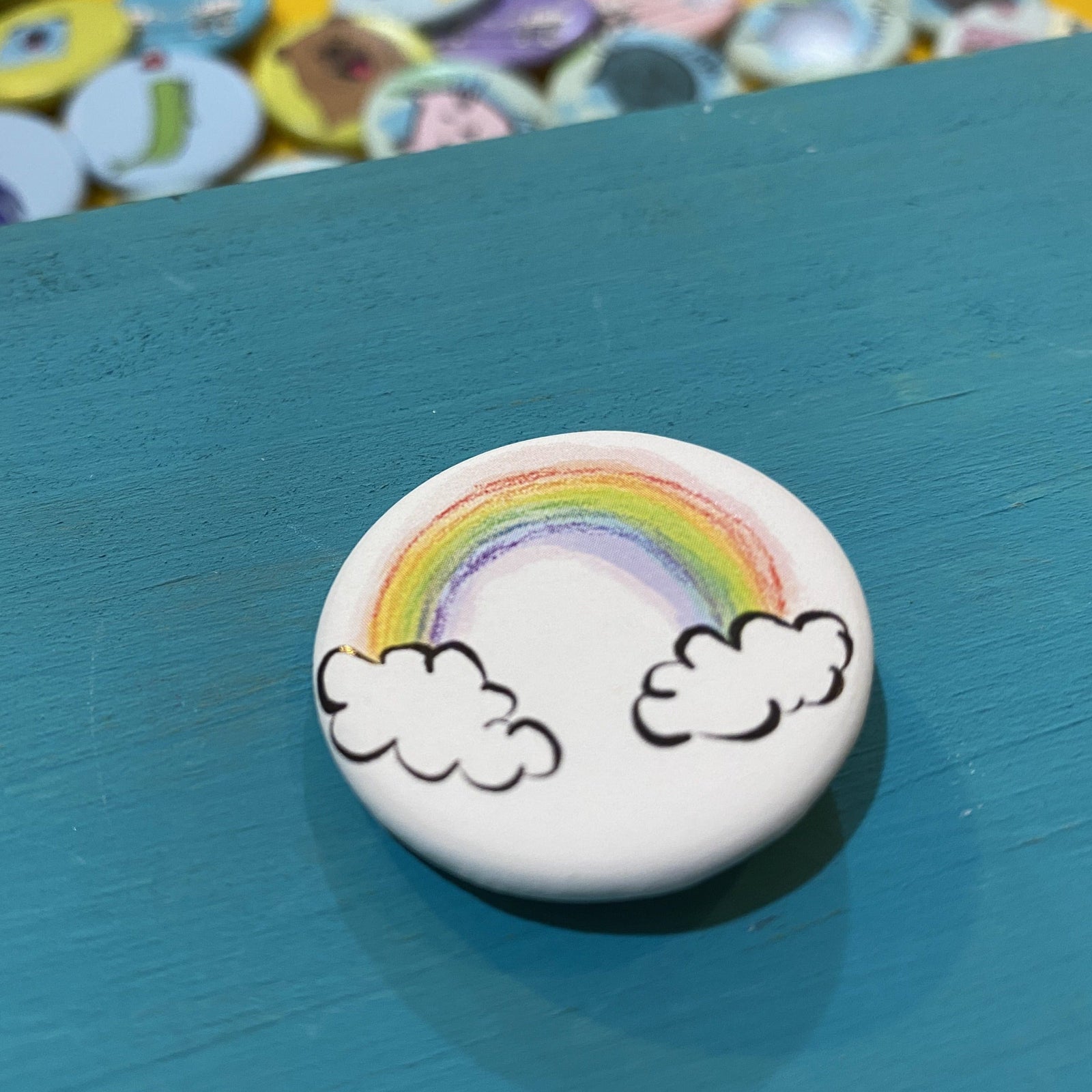 Button badge with a rainbow and clouds design on a blue surface