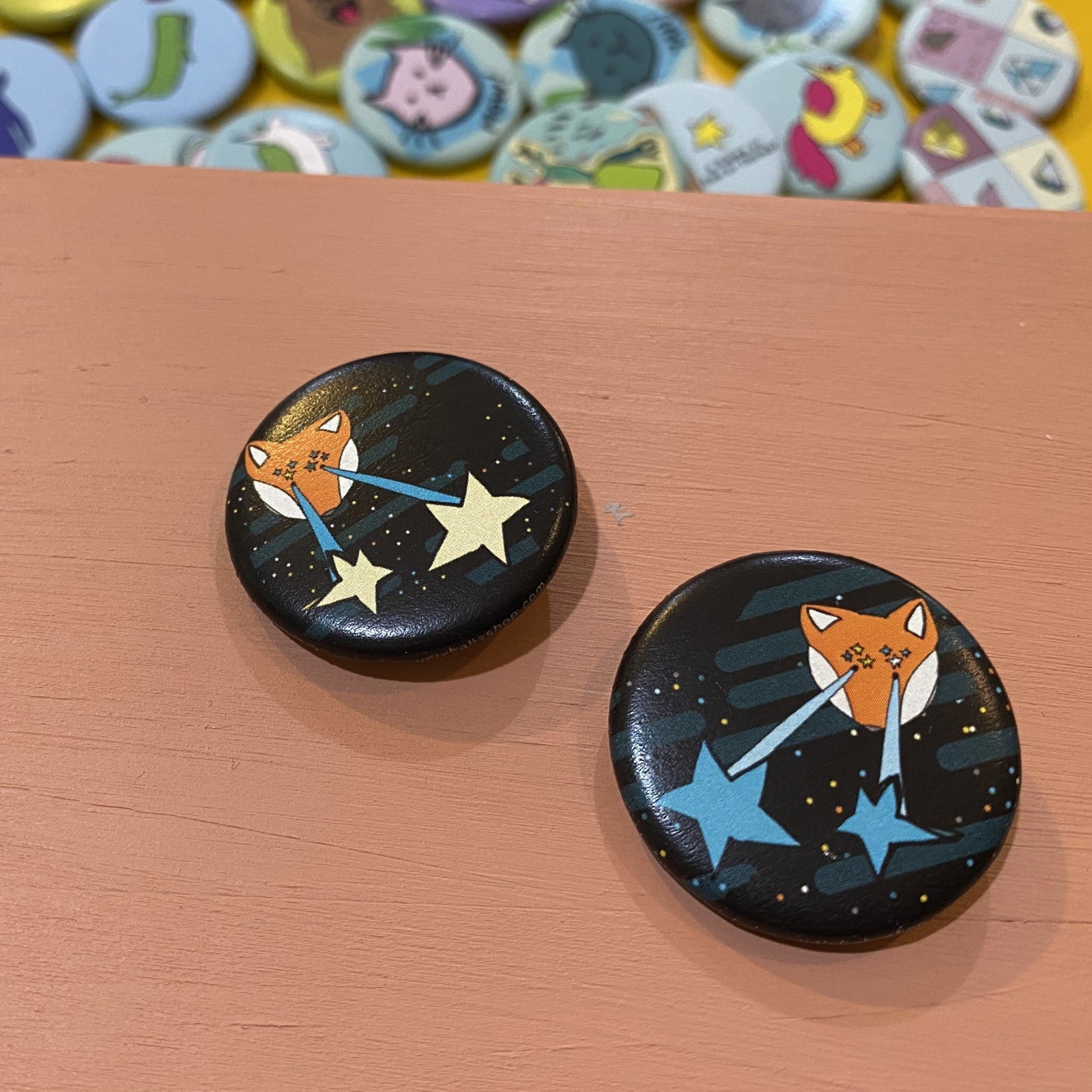 Two button pins with fox designs on a starry background, placed on a wooden surface.