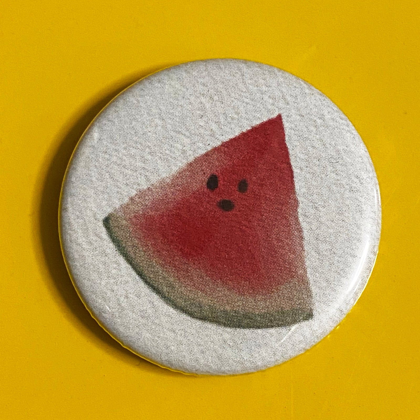A round, white button with a watercolor illustration of a watermelon on it, against a yellow background. 