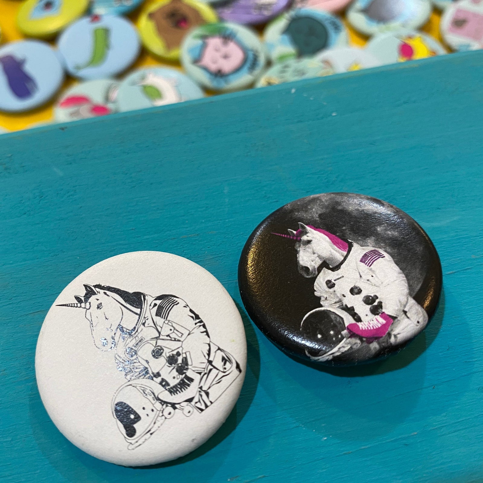 Two buttons with unicorn illustrations on a teal surface, with a colorful assortment of buttons in the background.
