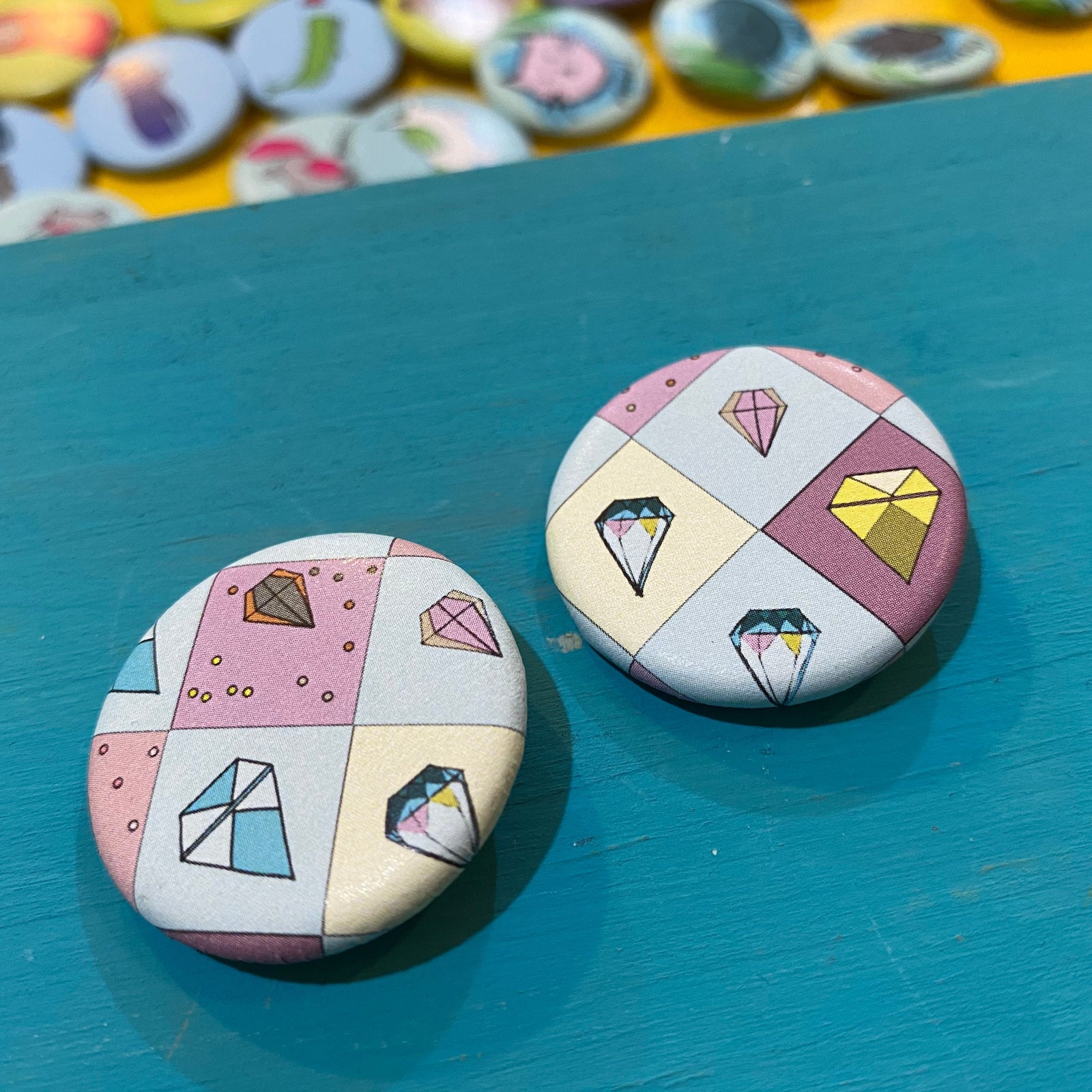 Two round pins with diamond pattern on a blue surface, with more pins in the background.
