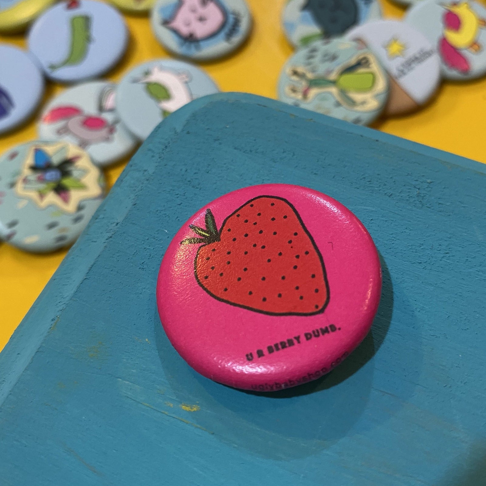 Pink button with a strawberry design and 'U R BERRY DUMB' text on a blue surface, with more buttons in the background.