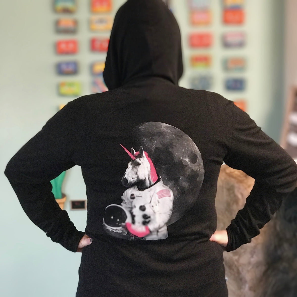 Black hooded sweatshirt with a roller skating unicorn astronaut screenprinted on the back in white and hot pink.  A person models with the hood up and their hands on their hips.