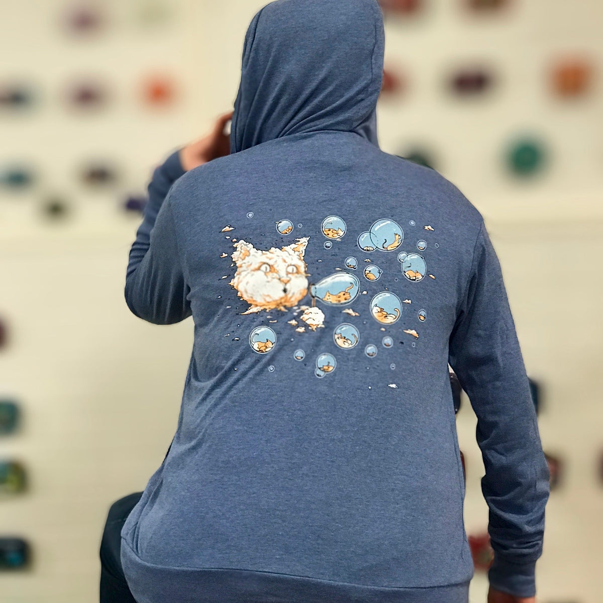 A light heathered blue hooded sweatshirt. On the back a cloud cat is blowing bubbles and inside the bubbles are sleeping kittens.