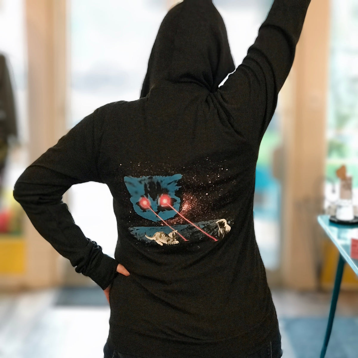 Heather black triblend unisex hooded sweatshirt. Giant space cat shooting pink lasers from his eyes. Tiny kittens are playing in the lasers on the surface of the moon.