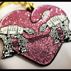 A closeup of a glittery pink keychain with two robots surrounded by a heart against a white background. 