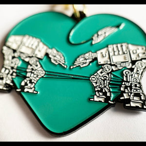 A closeup of a teal keychain featuring to robots surrounded by a heart against a white background. 