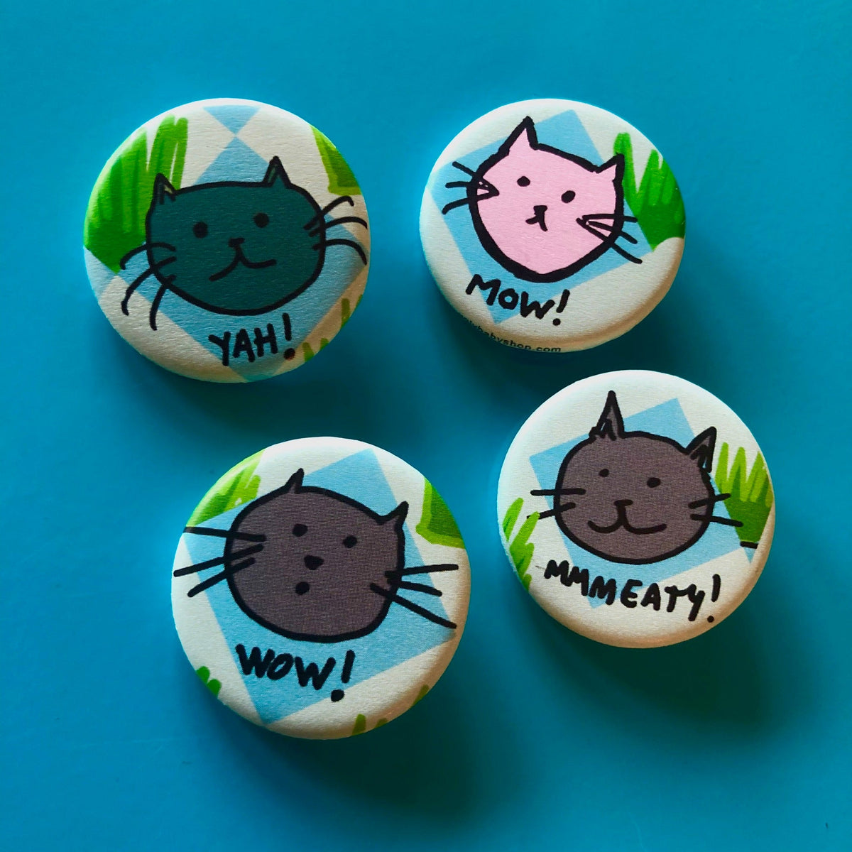 Four round magnets with cat face illustrations and text that says "yah!" "mow!" "wow!" and "mmmeaty!" against a blue background. 