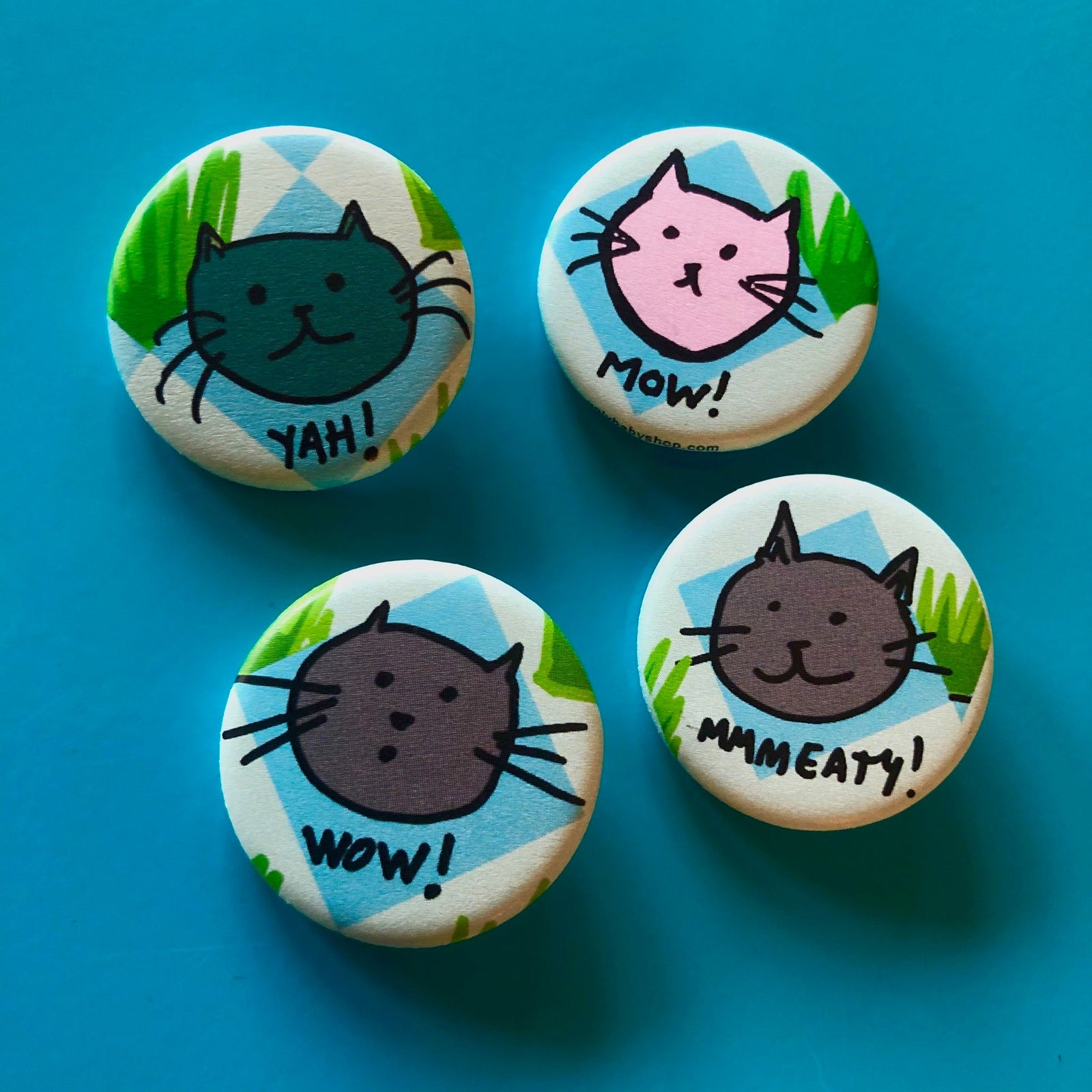 Four round magnets with cat face illustrations and text that says "yah!" "mow!" "wow!" and "mmmeaty!" against a blue background. 