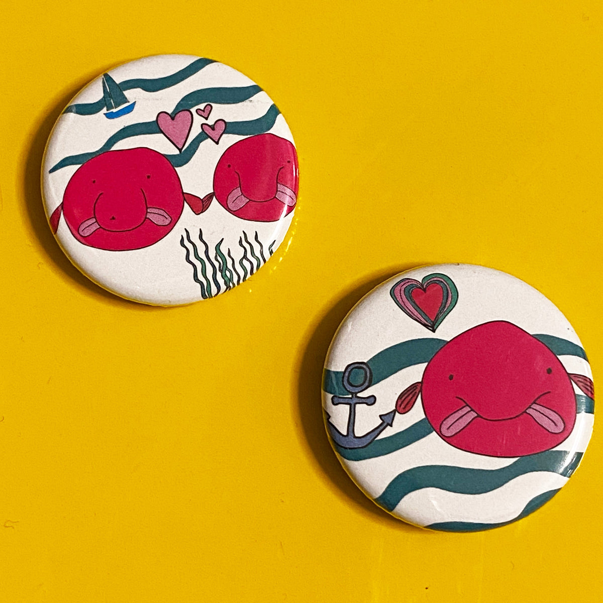 Two round magnets with illustrations of pink blobfish, hearts and anchors against a yellow background. 