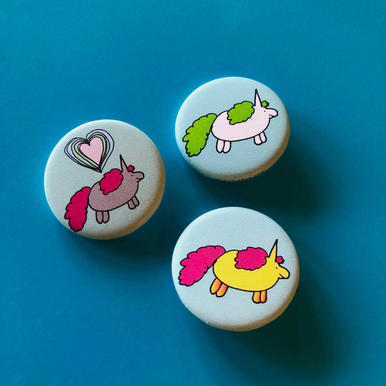 Three light blue round magnets with drawings of chubby unicorns on them against a blue background. 