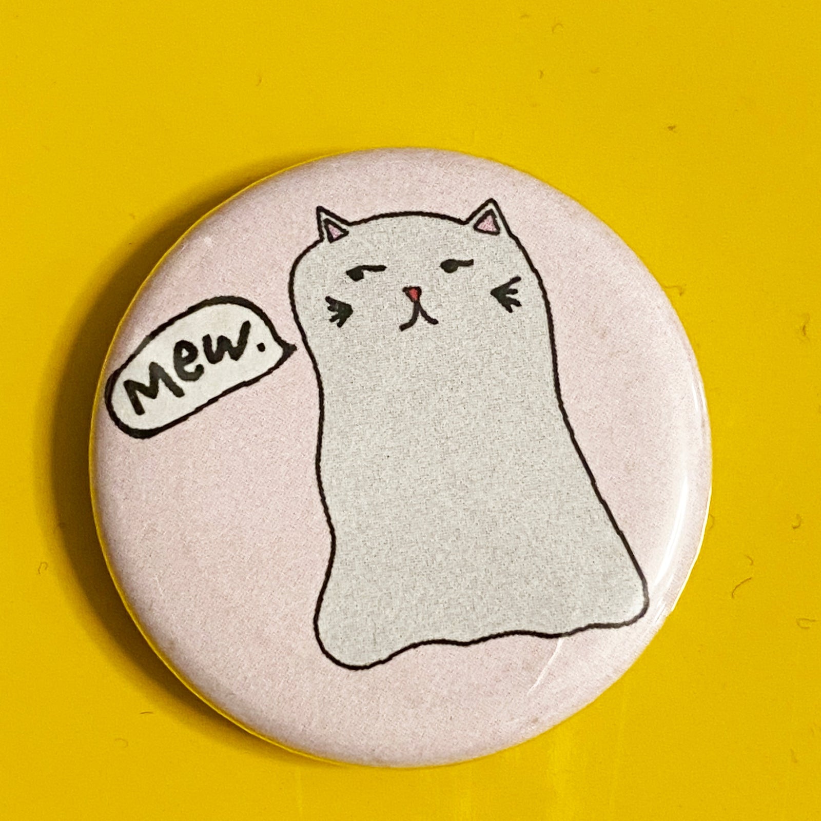A round pink magnet with an illustration of a ghost cat with a speech bubble sayign "mew." against a yellow background 