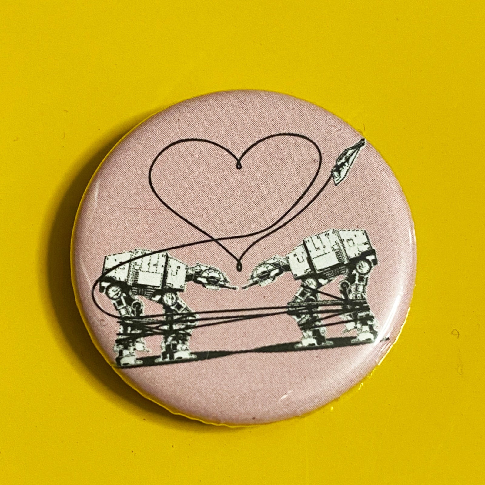 A round pink magnet with an illustration of two AT-ATs from Star Wars with a heart above them against a yellow background. 