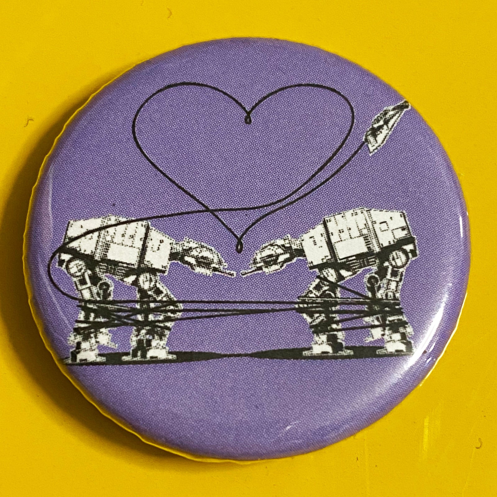 A round purple magnet with an illustration of two AT-ATs from Star Wars with a heart above them against a yellow background. 