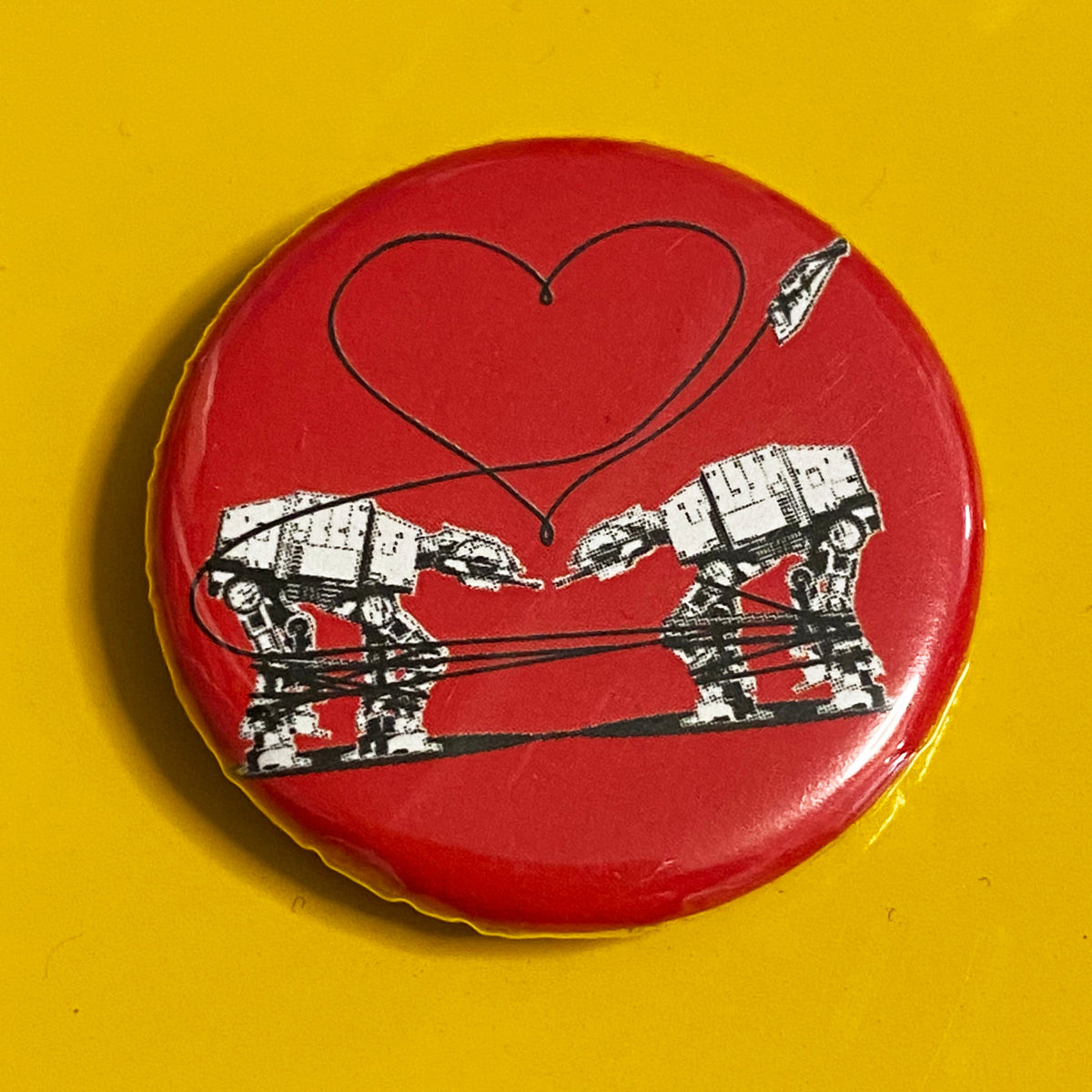 A round red magnet with an illustration of  two AT-ATs from Star Wars and a heart above them against a yellow background. 
