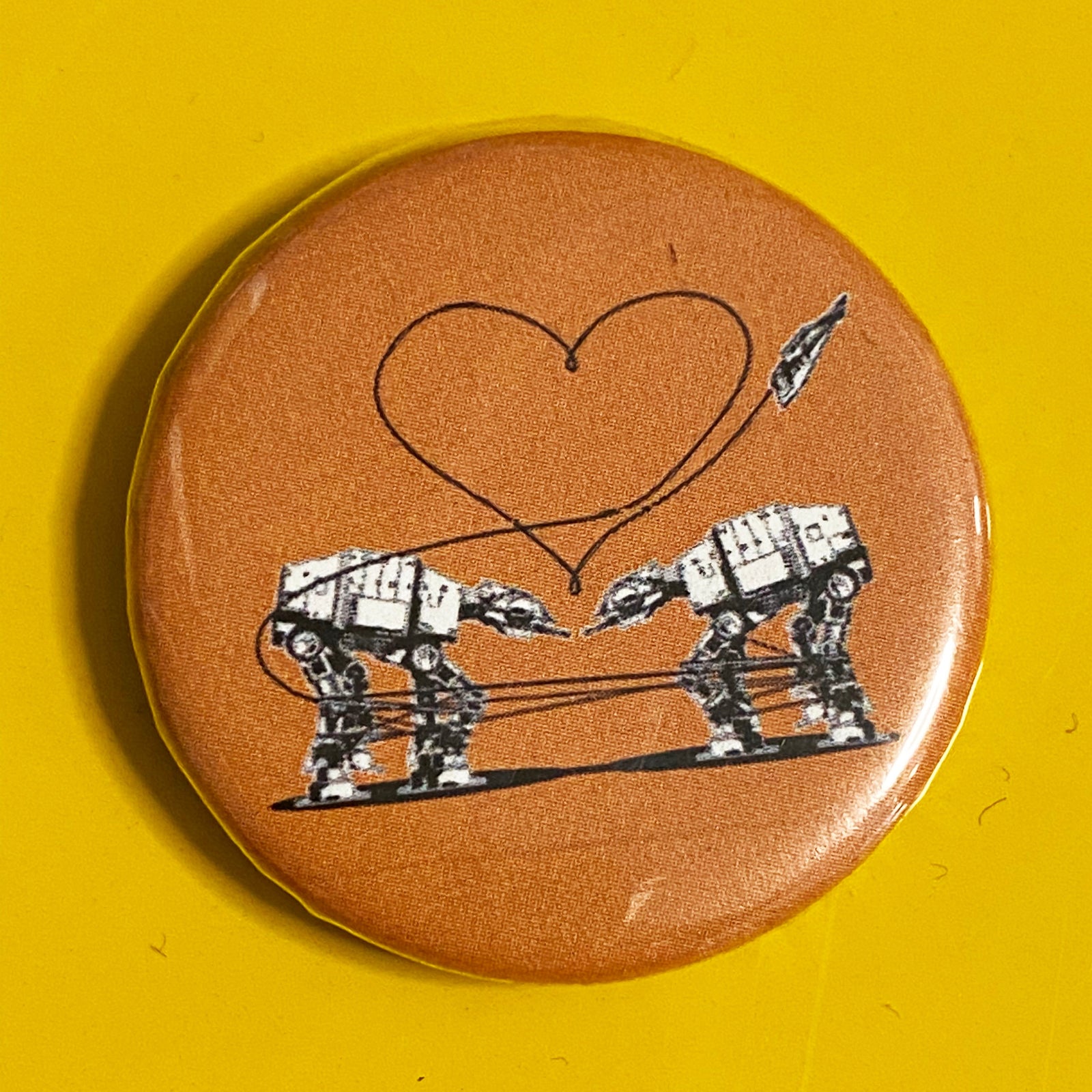 A round orange magnet featuring a design of two AT-ATs from Star Wars with a heart above them. 
