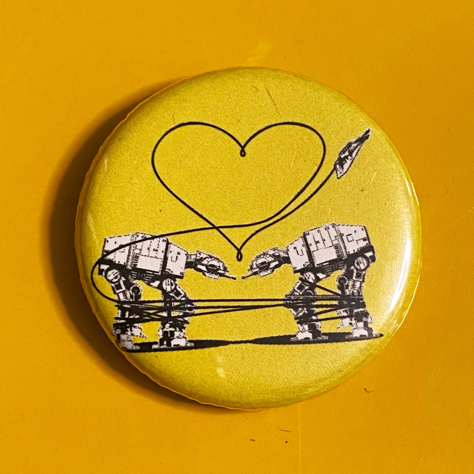 A yellow magnet with a design of two AT-ATs from Star Wars with a heart above them against a darker yellow background. 