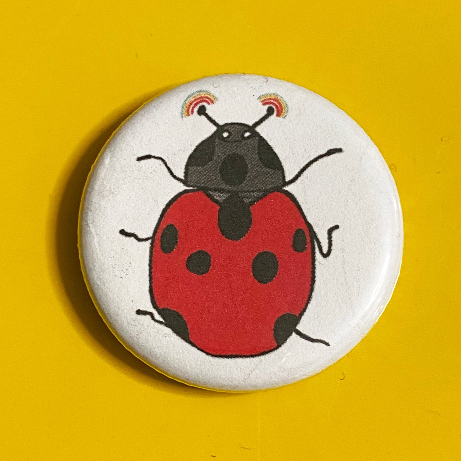 A round magnet with an illustration of a ladybug with rainbows shooting out of its antennae on it against a yellow backdrop. 