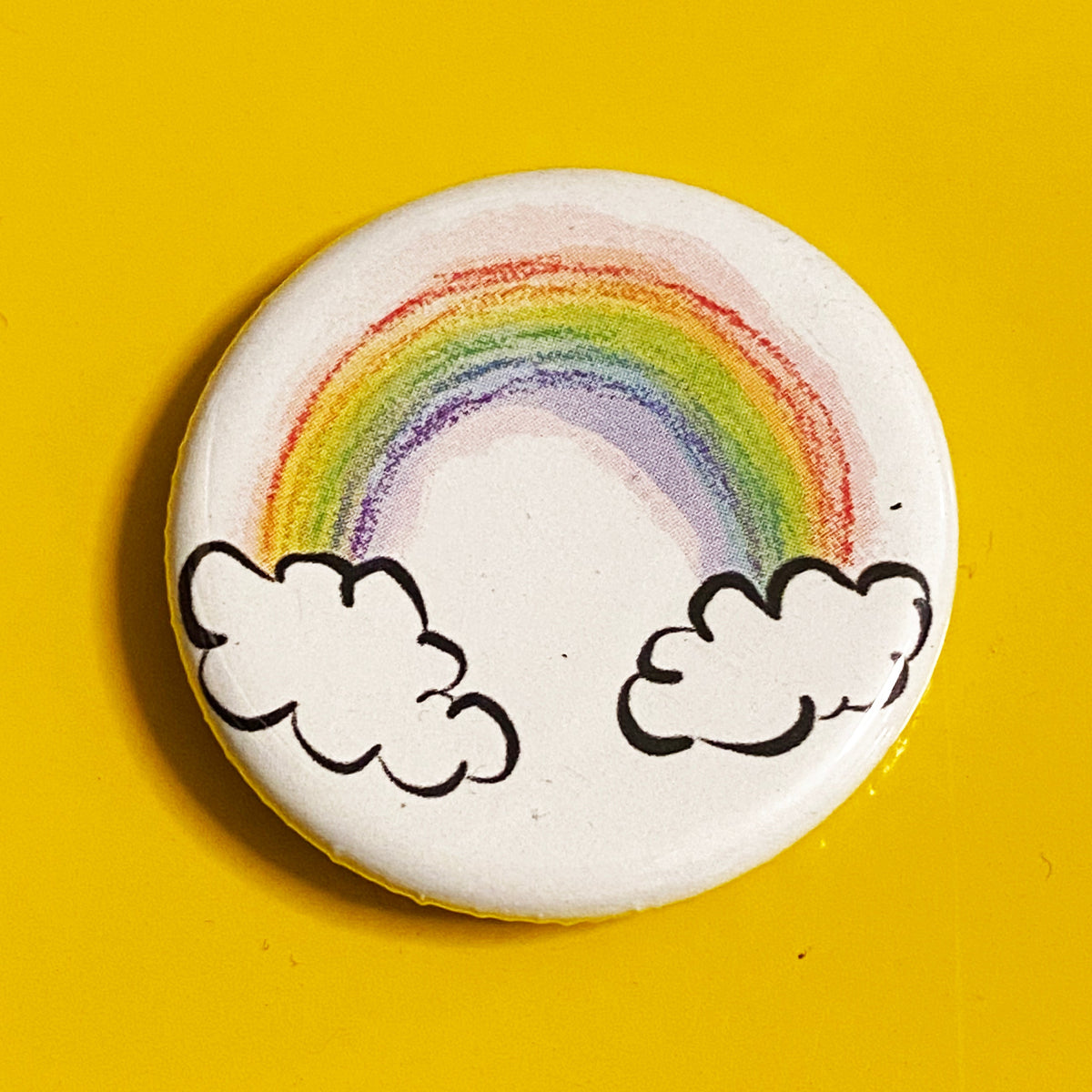 A round magnet with an illustration of a rainbow against a yellow background. 