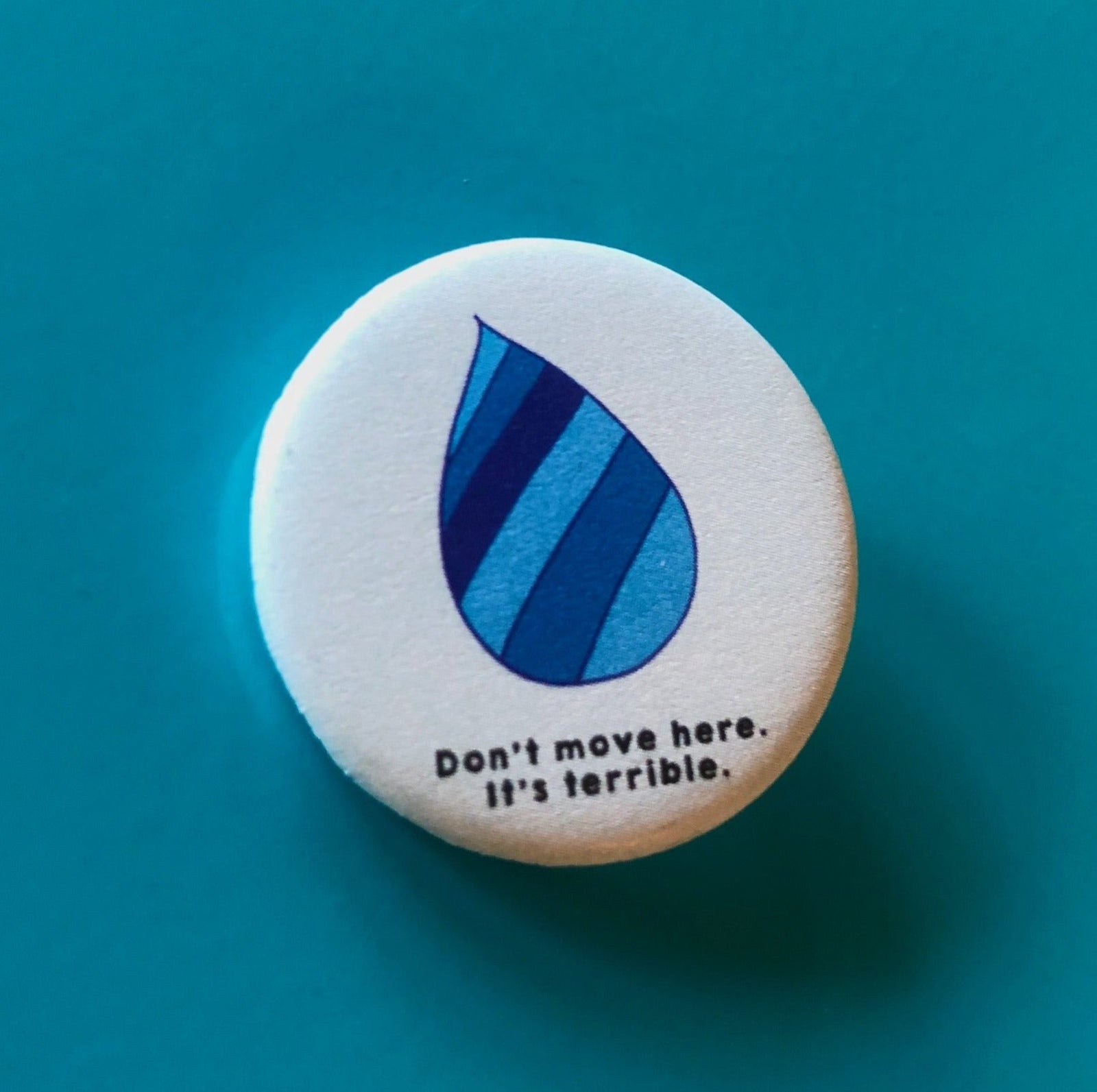 A round light blue magnet with a striped blue raindrop on it and text that says "Don't move here. It's terrible."